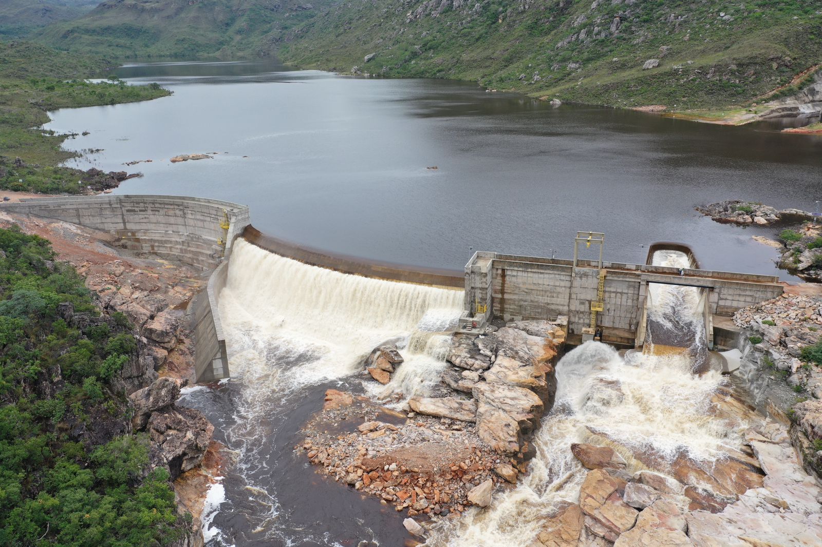 Barragem de Pequena Central Hidrelétrica (PCH) em área montanhosa, com reservatório cheio e água vertendo pelo vertedouro de concreto sobre rochas naturais.
