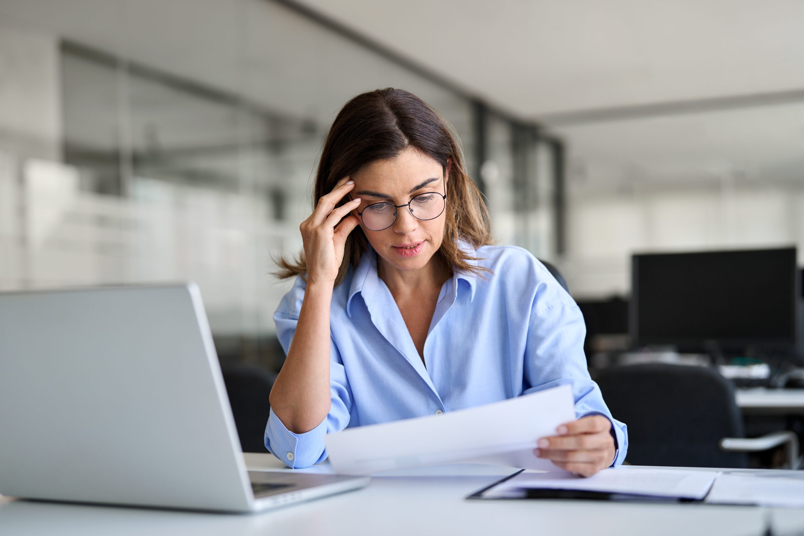 Mulher em ambiente corporativo segurando documentos, aparentando estar preocupada diante do notebook, simbolizando desafios profissionais e tomada de decisões.