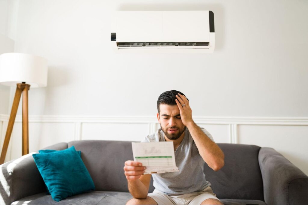 Homem sentado no sofá, segurando uma conta de energia e demonstrando preocupação, com um ar-condicionado instalado na parede ao fundo