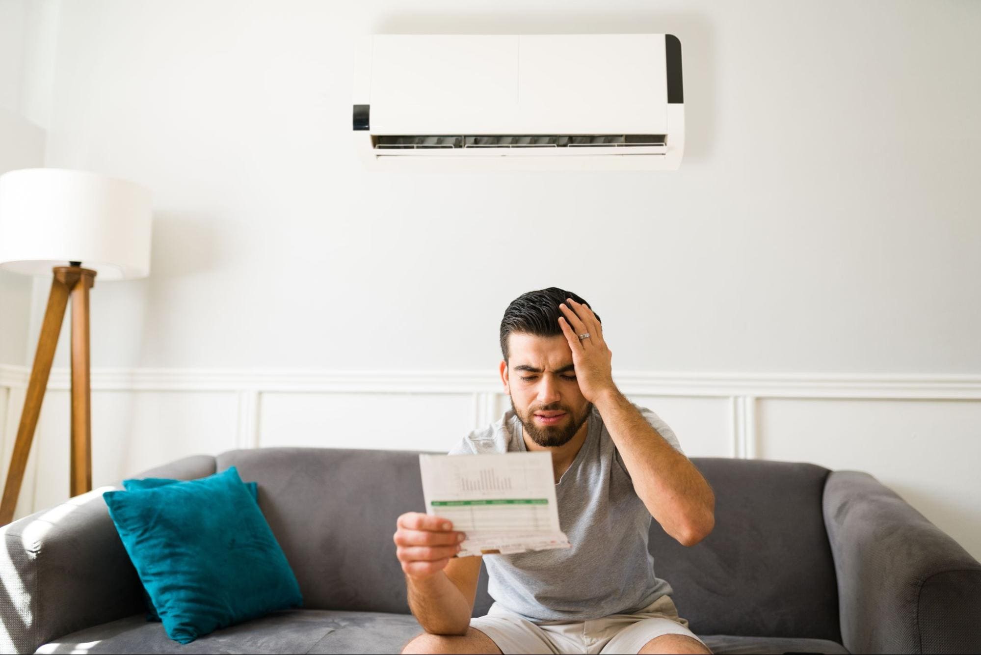 Homem sentado no sofá, segurando uma conta de energia e demonstrando preocupação, com um ar-condicionado instalado na parede ao fundo