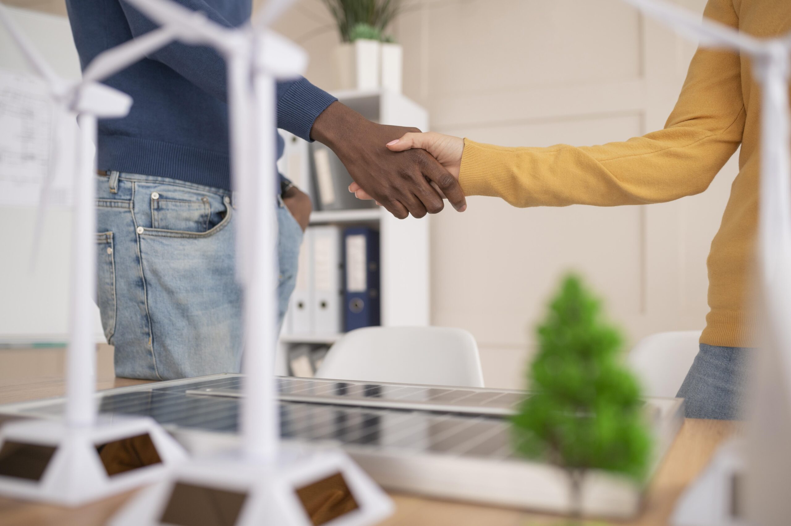 Homem e mulher apertam as mãos em sinal de acordo, com modelos de energia eólica e solar desfocados em primeiro plano.
