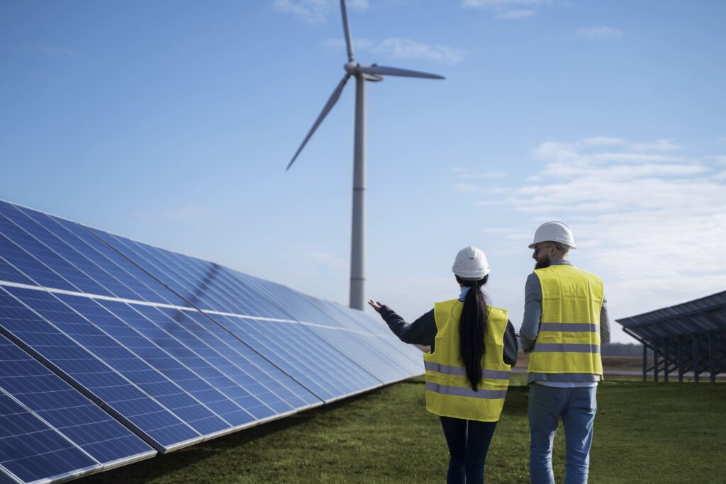 Dois profissionais usando capacetes e coletes refletivos analisam painéis solares em uma usina de geração distribuída, com uma turbina eólica ao fundo sob céu azu